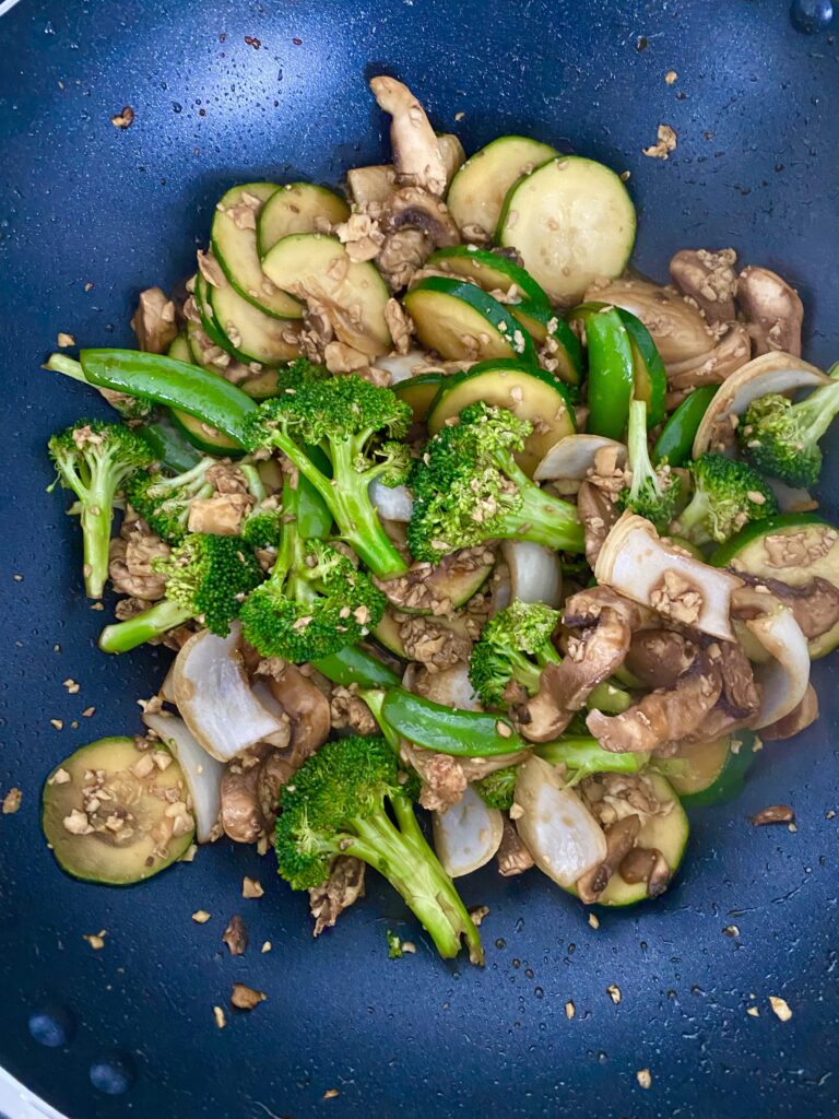 Green broccoli, green zucchini, green snap peas, brown/white mushrooms, white onions, and little chunks of white tofu all on a black pan